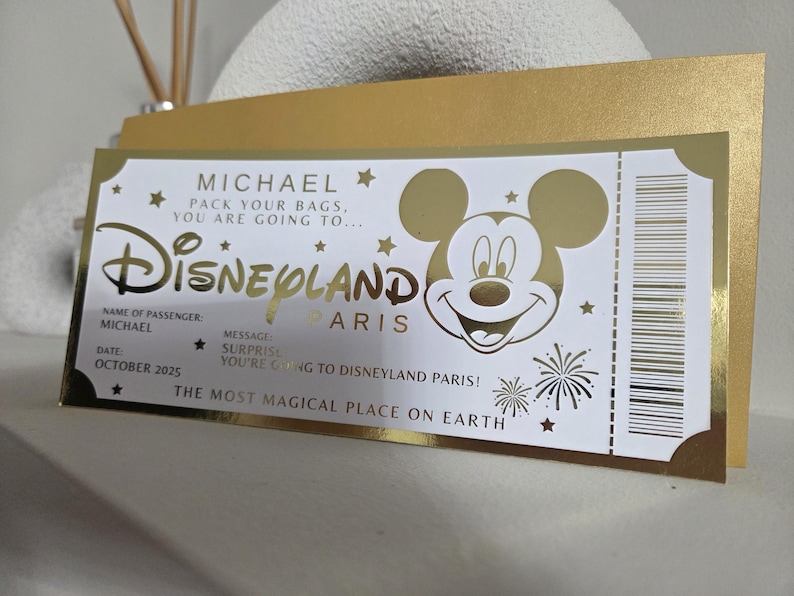 May include: A gold and white Disneyland Paris ticket-themed card. The card features the Disneyland logo, Mickey Mouse, and text that reads "MICHAEL, PACK YOUR BAGS, YOU ARE GOING TO..." and "THE MOST MAGICAL PLACE ON EARTH."