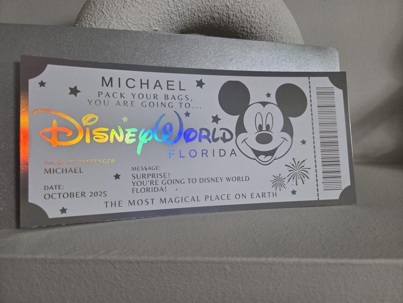 May include: A silver ticket-style card with the text "MICHAEL" and "Disney World Florida". The card features a Mickey Mouse graphic and the message "SURPRISE! You're going to Disney World Florida!" with the date "OCTOBER 2025".