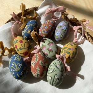 May include: Eight hand-painted Easter eggs with floral designs. The eggs are decorated with various flowers, including snowdrops, roses, and other spring blooms. The eggs are tied with pink and gold ribbons.
