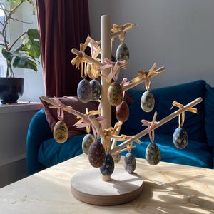 Peut inclure: Un arbre à œufs de Pâques en bois avec une base ronde, décoré d'œufs recouverts de tissu dans différents motifs floraux et couleurs, notamment jaune, bleu, vert, rose et marron. Les œufs sont attachés avec des rubans roses et jaunes.