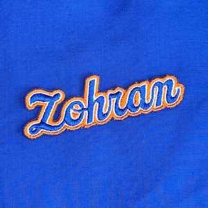 Może przedstawiać: Zbliżenie na królewski niebieski materiał z haftowaną naszywką. Naszywka przedstawia imię "Zohran" w kursywie. Litery są niebieskie z pomarańczowym obrysem.