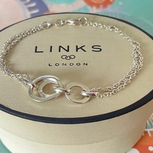 May include: A silver chain bracelet featuring two interlocking circular rings as a central focal point. The bracelet is displayed on a round, beige-coloured box with the black text LINKS LONDON printed on the top surface of the lid.