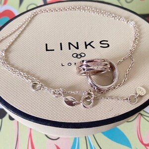 May include: A silver necklace with a pendant of interlocking rings, displayed on a beige box with the brand name "LINKS" printed in black. The necklace chain is delicate and features small, decorative links.