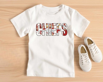 May include: White short-sleeved t-shirt with the word "CHIEFS" printed in black letters. The letters are made up of a collage of images of football players in red and white uniforms.