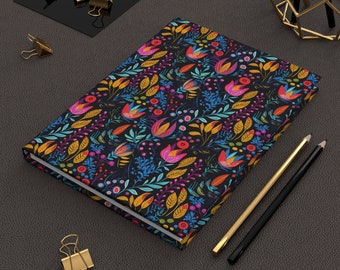 May include: A black notebook with a colorful floral pattern featuring red, yellow, orange, pink, blue, and green flowers and leaves. The notebook is lying on a dark gray surface with two pencils and gold paper clips.