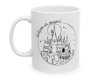 May include: White ceramic mug with a black and white illustration of a castle with a crescent moon and stars. The text "believe in magic" is written in a cursive font around the illustration.