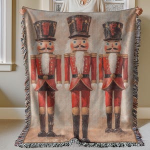 May include: A woven throw blanket with a vintage-inspired design featuring three toy nutcracker soldiers. The nutcrackers are dressed in red and gold uniforms with black hats and white beards. The background is a light beige with a distressed texture.