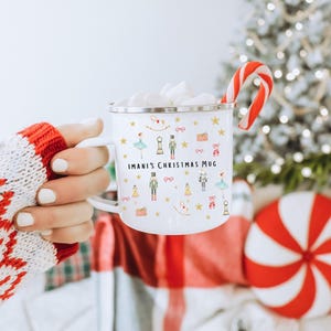 Puede incluir: Taza de cerámica blanca con borde y asa dorados. La taza está decorada con un diseño navideño festivo con un cascanueces, una bailarina y otros iconos navideños. El texto "IMANI'S CHRISTMAS MUG" está impreso en la taza.