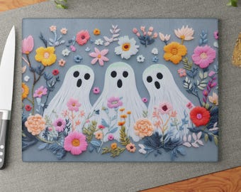 May include: A rectangular cutting board with a floral ghost design. Three white ghosts with black eyes and mouths are surrounded by colorful embroidered flowers in pink, yellow, and orange. The background is a muted blue-gray.
