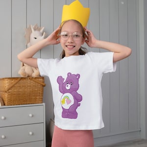 May include: A young person wearing a white t-shirt with a purple Care Bear with a rainbow and a heart on its belly. The person is wearing a yellow crown and glasses.
