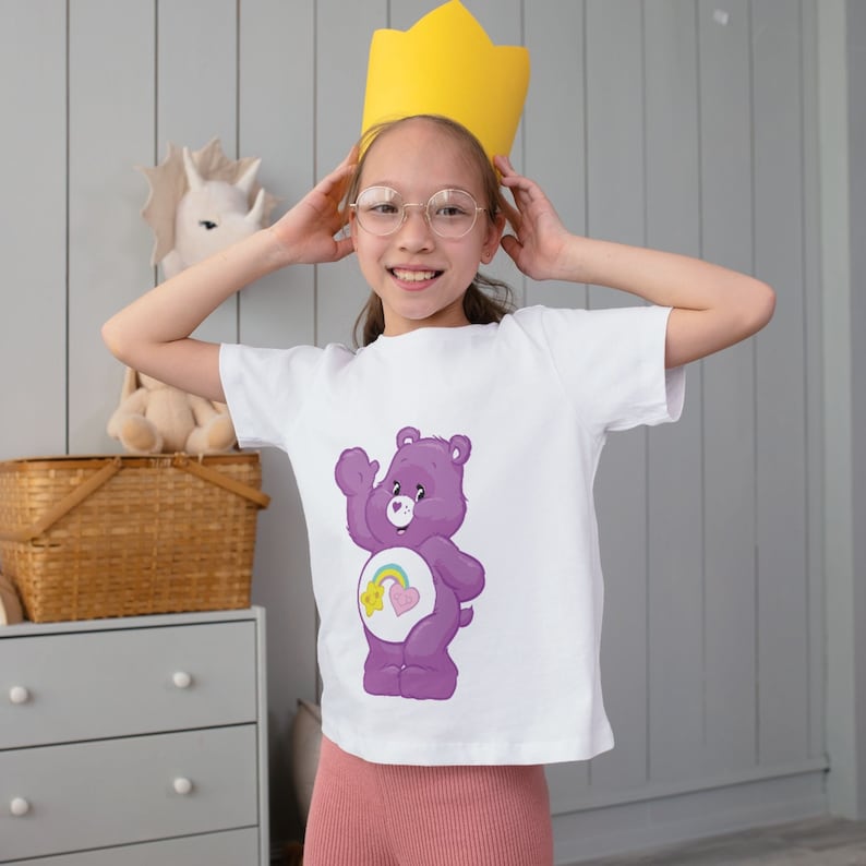 May include: A young person wearing a white t-shirt with a purple Care Bear with a rainbow and a heart on its belly. The person is wearing a yellow crown and glasses.