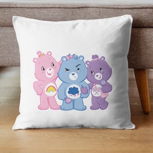 May include: Three Care Bears, a pink bear with a rainbow, a blue bear with a cloud, and a purple bear with a heart, are standing together on a white pillow.