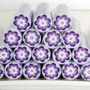 May include: A stack of polymer clay canes, each with a purple and white floral design, are arranged on a white rectangular dish with a gold rim. A U.S. dime is next to the dish. The canes are wrapped in clear plastic.