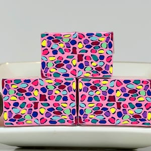 May include: Three pink, rectangular boxes with a colorful, abstract pattern of blue, yellow, purple, and green shapes. The boxes are on a white and pink dish, with a quarter coin for size comparison.