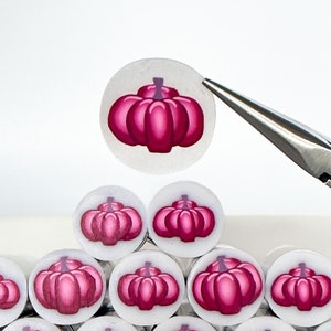 May include: A collection of small, round, white pumpkin-themed embellishments with a vibrant magenta color. The pumpkins are stacked in a pyramid shape, with one being held by silver tweezers. The embellishments are in a white dish.