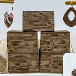 May include: An assortment of handcrafted earrings in various styles, including hoop earrings with pink and white accents, teardrop-shaped earrings, and small cross stud earrings. The earrings are displayed with wooden block props and a gold-colored stand.