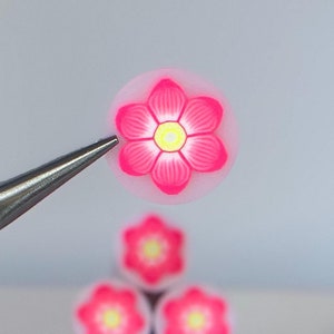 May include: Close-up of a pink flower bead held by tweezers, with a stack of similar beads in the background. The flower beads have a yellow center and are on a white surface. A single pink petal is also visible.