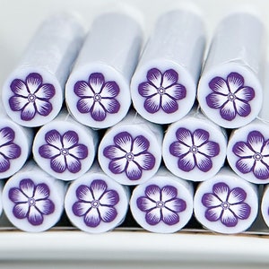May include: A stack of white polymer clay canes with a purple flower design on a white plate. The canes are cylindrical and have a flower pattern on the end. A dime is next to the plate for size comparison.
