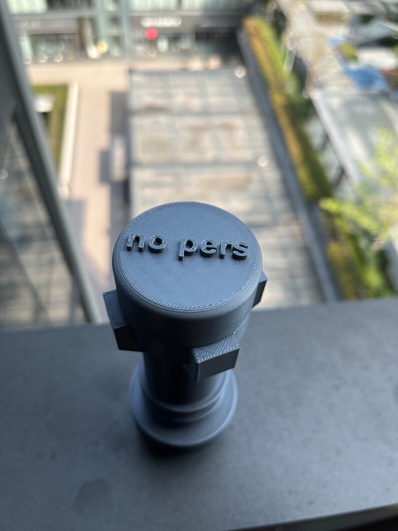 May include: A gray 3D printed object with the words "no pers" on top. The object has a cylindrical shape with a square base.