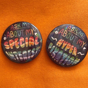 May include: Two round black buttons with rainbow text. The first button says "Ask me about my special interest". The second button says "Ask me about my hyper fixation".