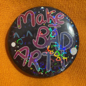 May include: A black button with a holographic finish features the text "Make Bad Art" in pink, blue, and purple lettering. The button is covered in a rainbow glitter pattern with small white hearts.