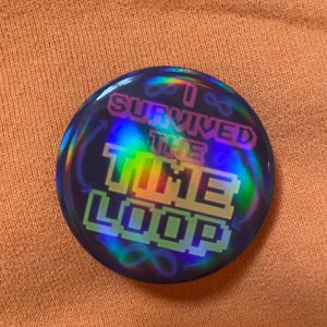 May include: A circular badge with a holographic effect. The text "I SURVIVED THE TIME LOOP" is displayed in a pixelated font, with infinity symbols. The badge has a dark blue background with rainbow hues.
