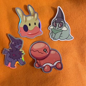 Puede incluir: Cuatro pegatinas holográficas con personajes de dibujos animados. Las pegatinas están sobre una superficie naranja brillante. Los personajes son una criatura rosa y amarilla parecida a una babosa, una criatura verde con una aleta morada, una criatura morada con una melena negra y una criatura roja con una barriga blanca.