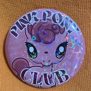 May include: Pink and purple holographic button with a cartoon pony with a pink and purple mane and tail. The button says "Pink Pony Club" in a pink and purple font.