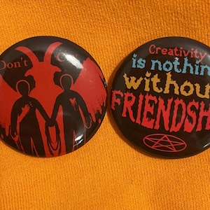 Puede incluir: Dos pines redondos negros con texto de colores. El primer pin presenta un diablo rojo con dos figuras de pie a cada lado. El texto en el pin dice "Don't". El segundo pin presenta el texto "Creativity is nothing without FRIENDSHIP" en un arcoíris de colores. El pin también presenta un pentagrama negro.