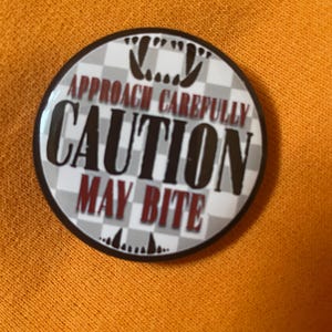 Puede incluir: Un botón redondo con el texto "ACÉRCATE CON CUIDADO PRECAUCIÓN PUEDE MORDER" en letras negras y rojas. El botón tiene un fondo a cuadros y un borde negro. Está sobre una superficie naranja.