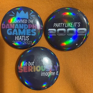 May include: Three round, holographic buttons with black backgrounds and colorful rainbow accents. The first button says "I survived the Dan and Phil Games Hiatus 2018-2023". The second button says "Party Like It's 2009". The third button says "No but Seriously imagine it".
