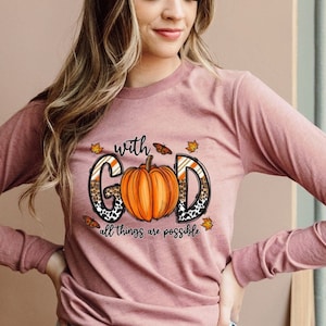 May include: Mauve long-sleeve shirt featuring a pumpkin graphic with the words "with God all things are possible." The pumpkin is orange with a leopard print design. Autumn leaves surround the text and pumpkin.