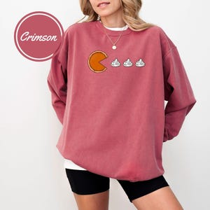 May include: A crimson sweatshirt with a graphic of a slice of pumpkin pie with a Pac-Man mouth eating three mini marshmallows. The text "Crimson" is in a circle on the left side of the image.