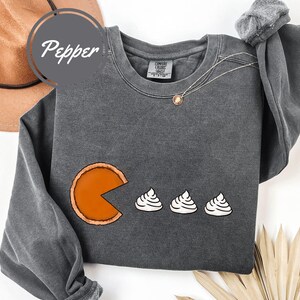 May include: A dark gray sweatshirt with a graphic of a pumpkin pie slice eating three mini pumpkin pies. The text "Comfort Colors" is visible on the tag.