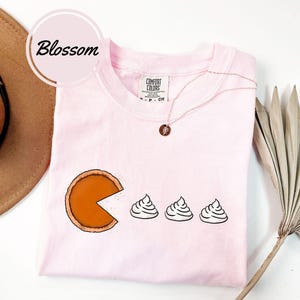 May include: Pink t-shirt with a graphic of a pumpkin pie with a bite taken out of it and three dollops of whipped cream. The text "Comfort Colors" is on the tag.
