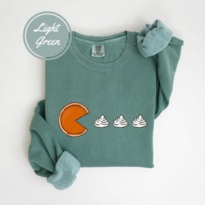May include: A light green sweatshirt with a graphic of a pumpkin pie slice eating three mini marshmallows. The text "Comfy Colors" is visible on the shirt.