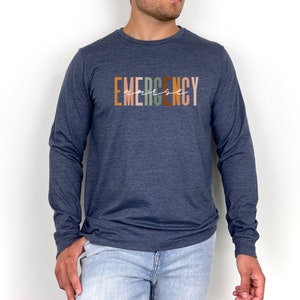 May include: A navy blue long-sleeved shirt with the text "EMERGENCY" printed in colorful letters. The word "arise" is written in white script underneath the word "EMERGENCY".
