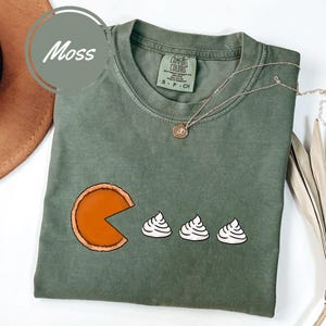 May include: A green t-shirt with a graphic of a pumpkin pie with a bite taken out of it and three dollops of whipped cream. The shirt has the text "COMFORT COLORS" and "S - P - CH" on the tag.