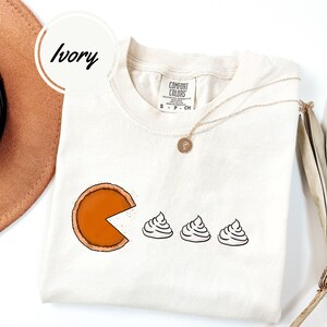May include: A white t-shirt with a graphic of a pumpkin pie with a slice missing and three dollops of whipped cream. The shirt has the text "Comfort Colors" on the tag.