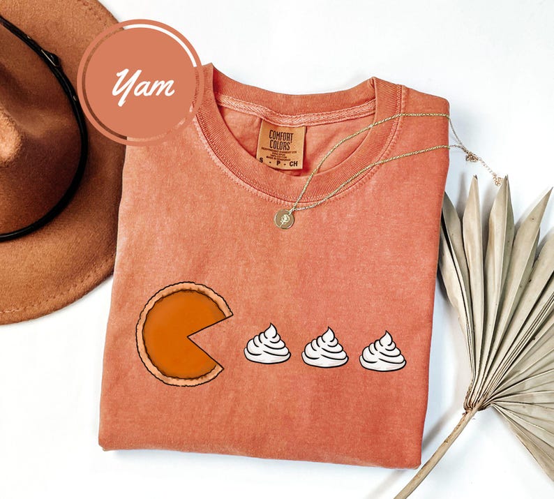 May include: A burnt orange t-shirt with a graphic of a pumpkin pie with a bite taken out of it and three dollops of whipped cream. The text "Comfort Colors" is visible on the shirt.
