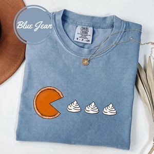 May include: A blue jean t-shirt with a graphic of a pumpkin pie slice with three mini pies on a blue background. The shirt has the text "Comfort Colors" and "S - P - CH" on the front.