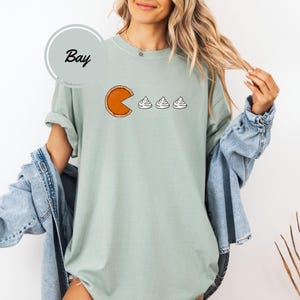 May include: A light green t-shirt with a graphic of a pumpkin pie with a bite taken out of it, chasing after three mini marshmallows. The text "Bay" is on a white circle in the top left corner of the image.