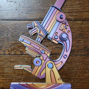 May include: A 3D printed model of a microscope in shades of purple, orange, and gold. The microscope is detailed with intricate lines and patterns.
