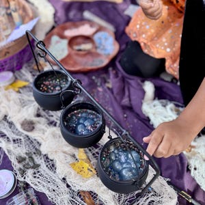 Peut inclure: Trois petits chaudrons noirs suspendus à un support métallique, remplis d'objets bleus et irisés. Les chaudrons sont posés sur une surface violette avec un voile blanc. D'autres bocaux et objets décoratifs sont également visibles.