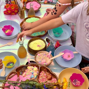 May include: A sensory play station featuring wooden divided trays filled with colorful sand, small beads, and textured powders. The setup includes light blue and yellow bowls, glass vials with liquids, wooden spoons, and fresh flowers.