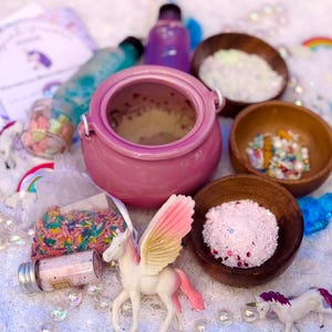 May include: A pink ceramic cauldron surrounded by wooden bowls containing white and pink powders, colorful sprinkles, and small bottles. A white unicorn figurine with pink and gold wings stands in front, alongside a bag of rainbow rice.