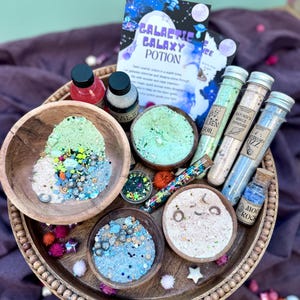 May include: A wooden tray displays a "Galactic Galaxy Potion" kit. It features small wooden bowls with colourful powders and glitter, glass tubes, and small bottles. A booklet labelled "Galactic Galaxy Potion" is also included, offering a magical theme.