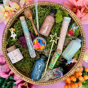 May include: A wooden bowl filled with fairy-themed bath products, including bath salts, bath bombs, and glitter. The bowl is surrounded by pink flowers and green moss.