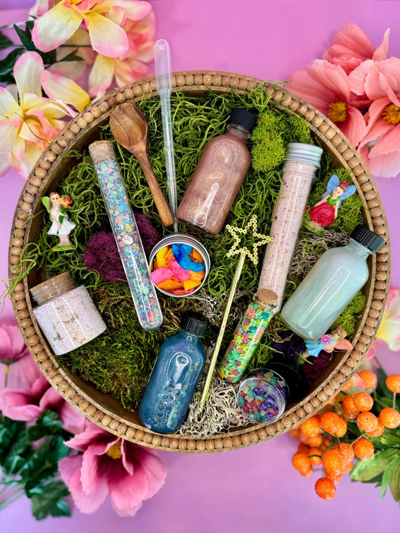 May include: A wooden bowl filled with fairy-themed bath products, including bath salts, bath bombs, and glitter. The bowl is surrounded by pink flowers and green moss.