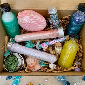 K&ouml;nnte beinhalten: Eine braune Pappbox gef&uuml;llt mit Bade- und K&ouml;rperprodukten, darunter eine rosa Muschel, eine Flasche mit gr&uuml;ner Fl&uuml;ssigkeit, eine Flasche mit blauer Fl&uuml;ssigkeit, eine Flasche mit gelber Fl&uuml;ssigkeit und mehrere R&ouml;hrchen mit Badesalz. Die Box ist umgeben von kleinen Dekorationsartikeln wie Muscheln, Perlen und Kristallen.